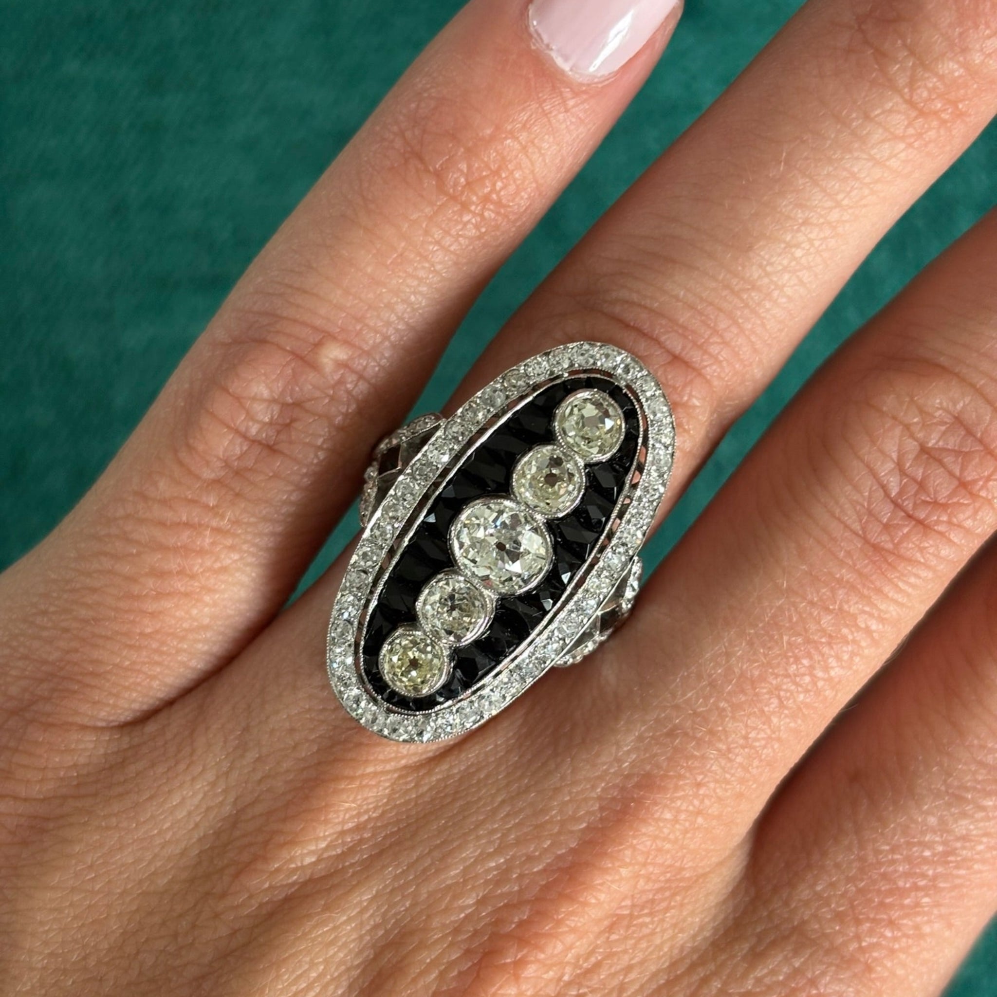 A hand with light skin wears the Onyx and Diamond Ring, an oval piece featuring five old mine-cut diamonds in a row, bordered by calibré-cut onyx and smaller diamonds, all crafted in platinum 1930s style.