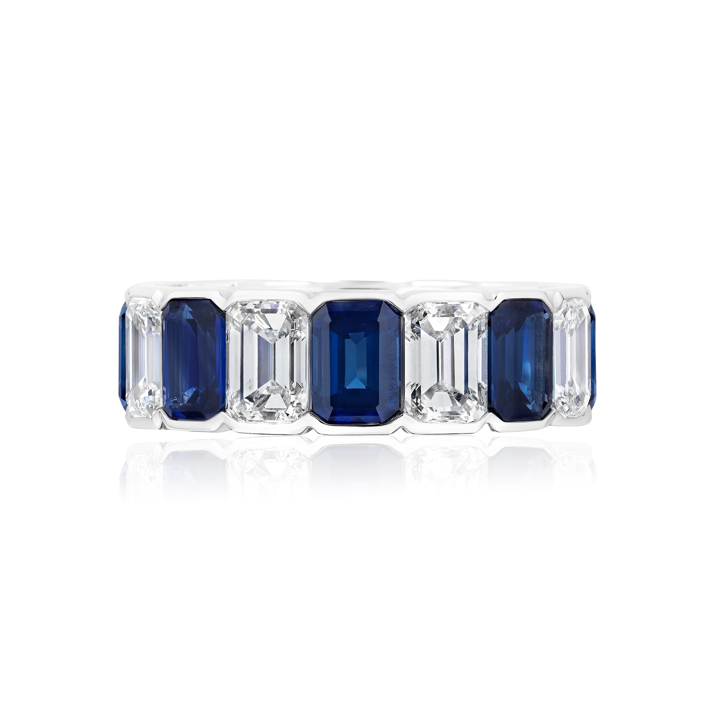 The Alternating Half-Bezel Emerald Cut Diamond and Sapphire Eternity Band showcases a white gold band with alternating emerald-cut blue sapphires and white diamonds, set in a continuous design and displayed on a white background.