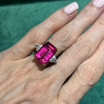 A hand with light pink nails wears the Pink Tourmaline and Diamond Ring, featuring an emerald-cut pink tourmaline and two triangular diamonds on a gold band, set against a green fabric background.