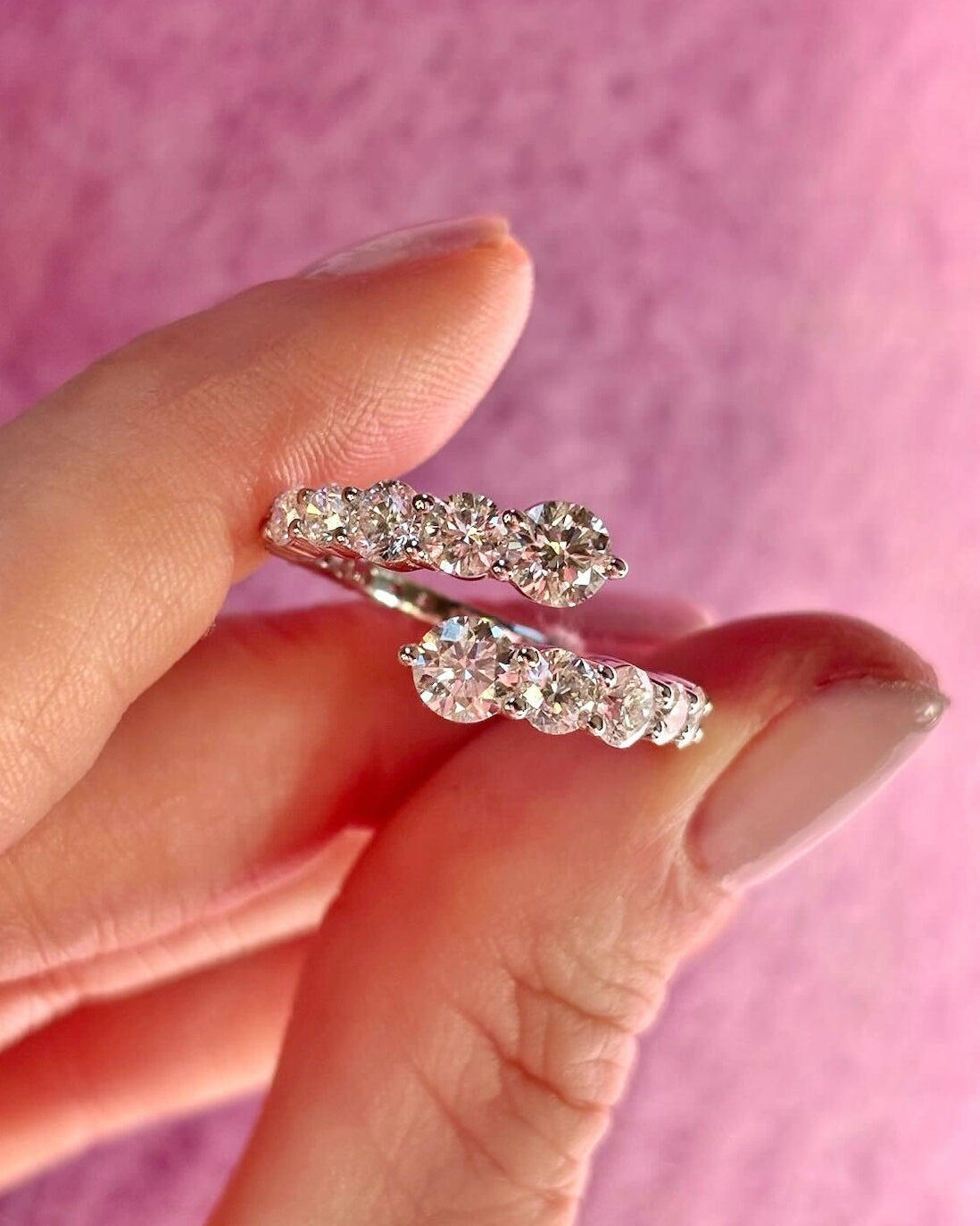 A manicured hand holds the Graduated Round Diamond Wraparound Ring in White Gold, featuring a row of sparkling round diamonds, against a soft pink background.