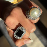 A hand displays the David Webb Pearl and Diamond Ring, showcasing a cultured pearl encircled by diamonds in 18K white gold under bright natural light, alongside a bold black gemstone ring with baguette-cut diamonds.