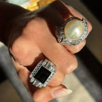 A hand adorned with two rings: the Onyx and Diamond Ring, featuring an emerald-cut onyx framed by diamonds in platinum, and a pearl ring encircled by diamonds. Sunlight highlights the dazzling jewels and graceful hand.