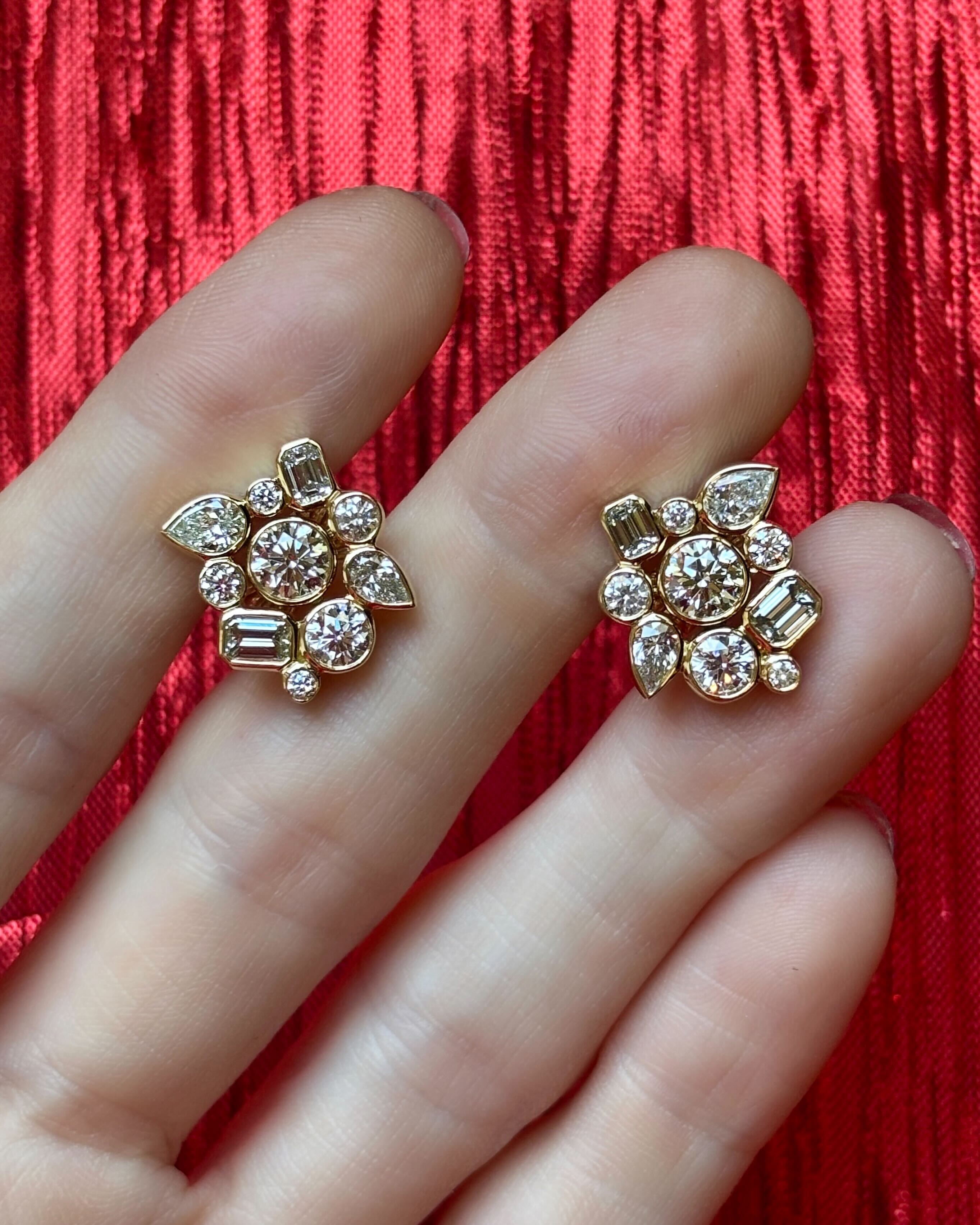 A pair of Bezel Set Mixed Shape Diamond Cluster Earrings in yellow gold are held between fingers against a red textured fabric, showcasing round, marquise, and baguette-cut diamonds set in elegant 18K gold.