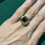 A hand is shown against a green background, wearing the 1970s Gold and Sapphire Ring—a gold ring featuring an elegant setting with a dark blue oval gemstone reminiscent of classic designs from the era.