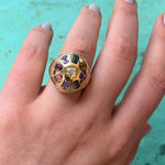 A hand with light-colored nail polish displays the Confetti Bombé Ring in 18k yellow gold, showcasing a central clear stone encircled by customizable multicolored gemstones, all set against a turquoise background.