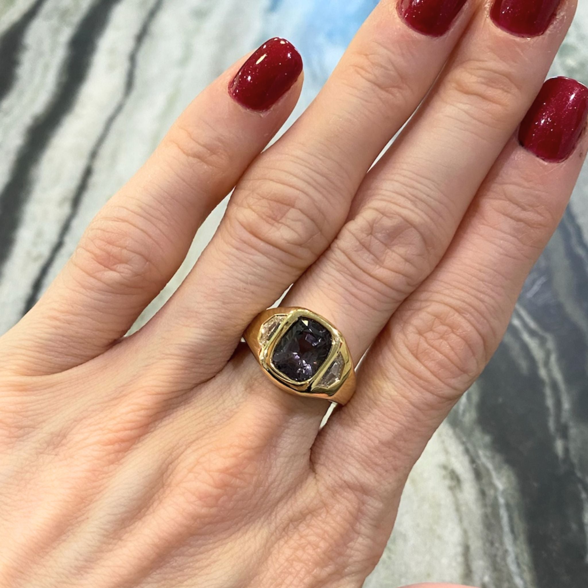A hand with red-painted nails wears the Cushion Cut Spinel and Diamond Epaulette Side Stone Gypsy Ring in Yellow Gold—an elegant 18k yellow gold ring showcasing a large cushion cut spinel accented by sparkling diamond side stones—displayed against a marbled surface.