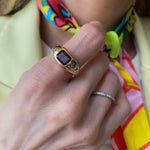 A hand adorned with the Bombé Ring, featuring an east-west set emerald cut ruby in yellow gold, as well as a thin silver band. The person wears a colorful scarf and a light yellow jacket.