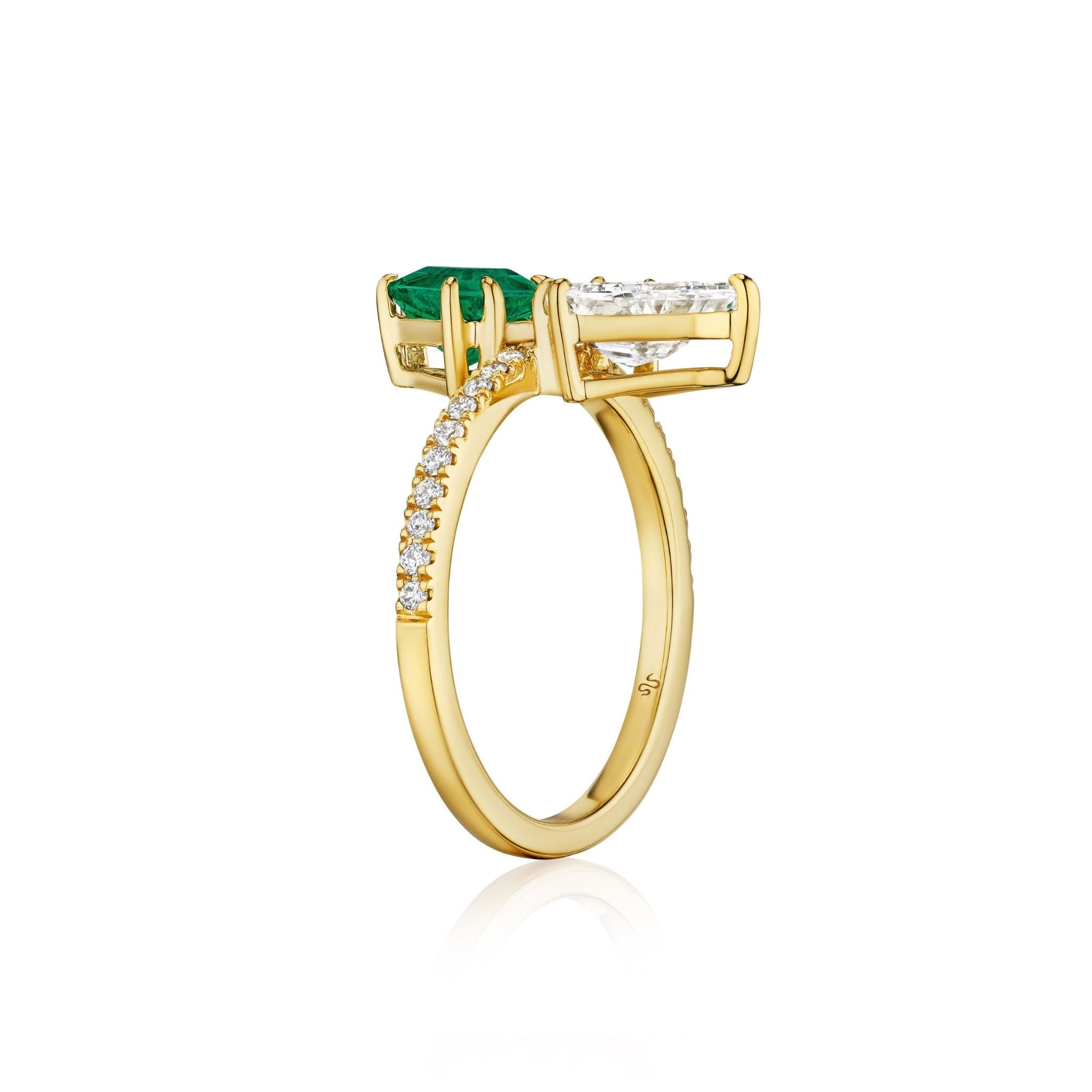 The Emerald and Diamond Double Shield Ring in Yellow Gold showcases a striking shield cut emerald and a dazzling shield cut diamond set side by side, with small diamonds adorning half of the 18k yellow gold band, pictured against a white background.