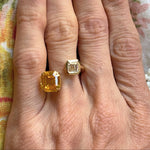 A hand with gold-painted nails displays the elegant Yellow Sapphire and Diamond Toi et Moi Ring in 18k yellow gold, featuring two large rectangular stones—an Asscher cut diamond and a vibrant yellow sapphire—set against a floral fabric background.