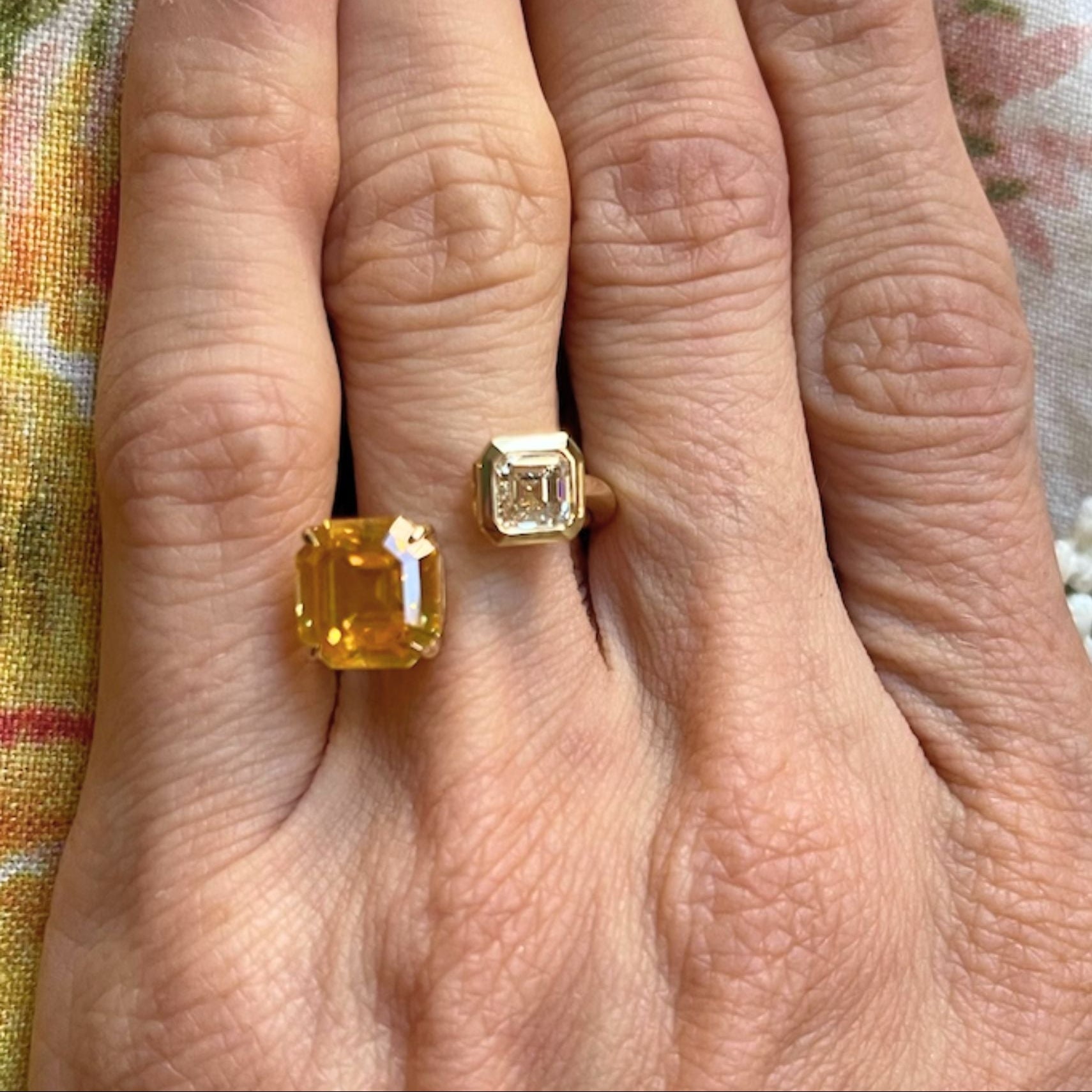 A hand with gold-painted nails displays the elegant Yellow Sapphire and Diamond Toi et Moi Ring in 18k yellow gold, featuring two large rectangular stones—an Asscher cut diamond and a vibrant yellow sapphire—set against a floral fabric background.