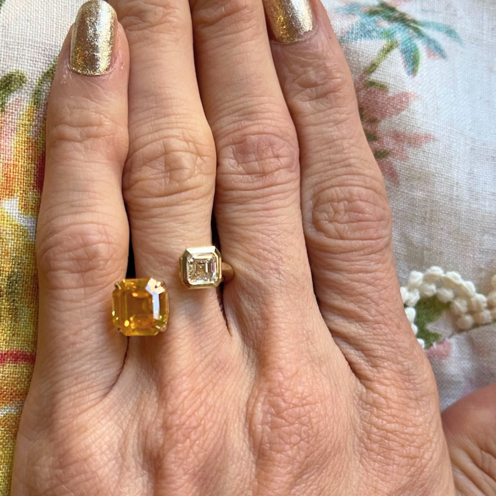 A hand with gold-painted nails displays the elegant Yellow Sapphire and Diamond Toi et Moi Ring in 18k yellow gold, featuring two large rectangular stones—an Asscher cut diamond and a vibrant yellow sapphire—set against a floral fabric background.