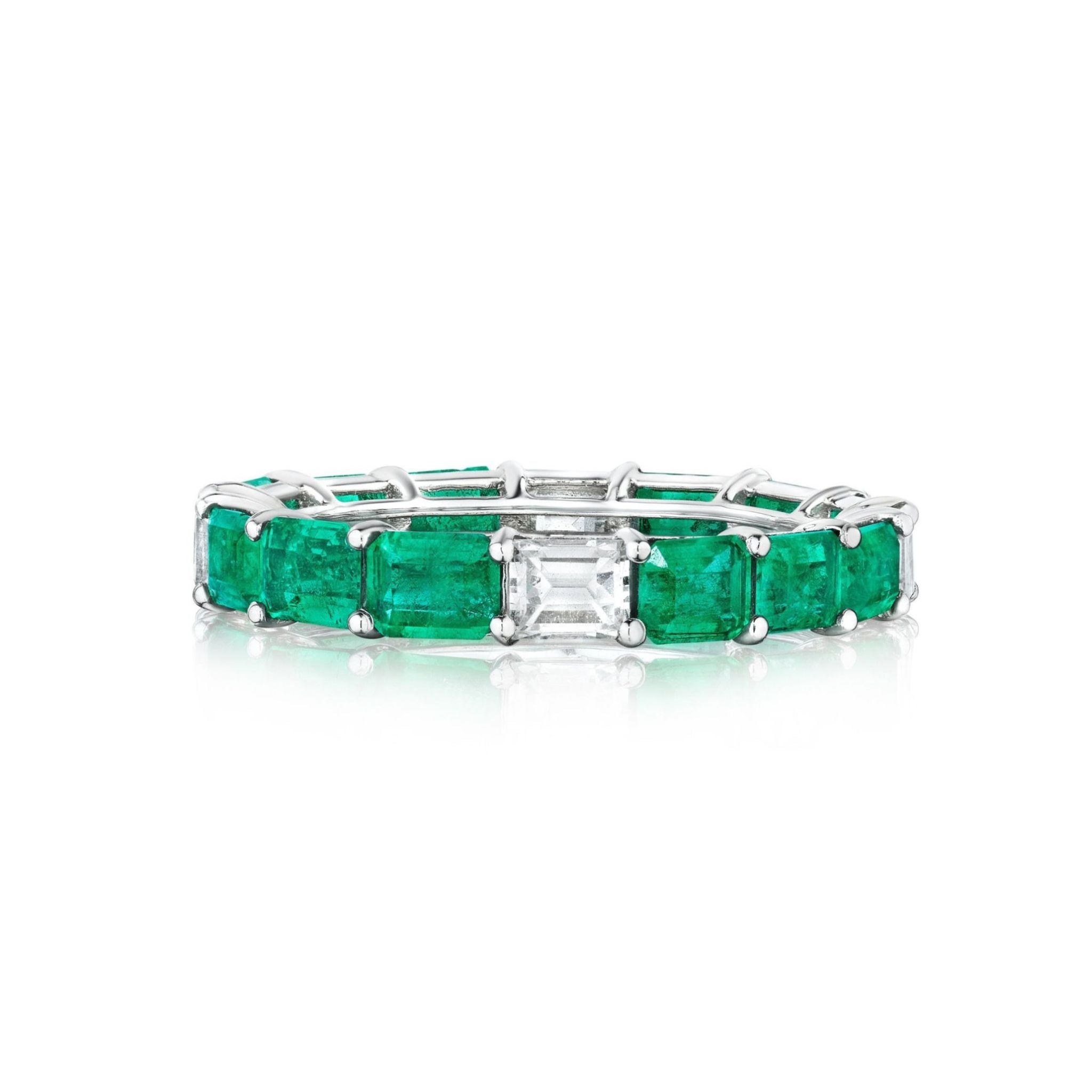 The East-West Emerald Cut Emerald and Diamond Eternity Band in White Gold is showcased on a white background, featuring an elegant row of emerald cut emeralds set in 18k white gold with a single rectangular diamond at the center.
