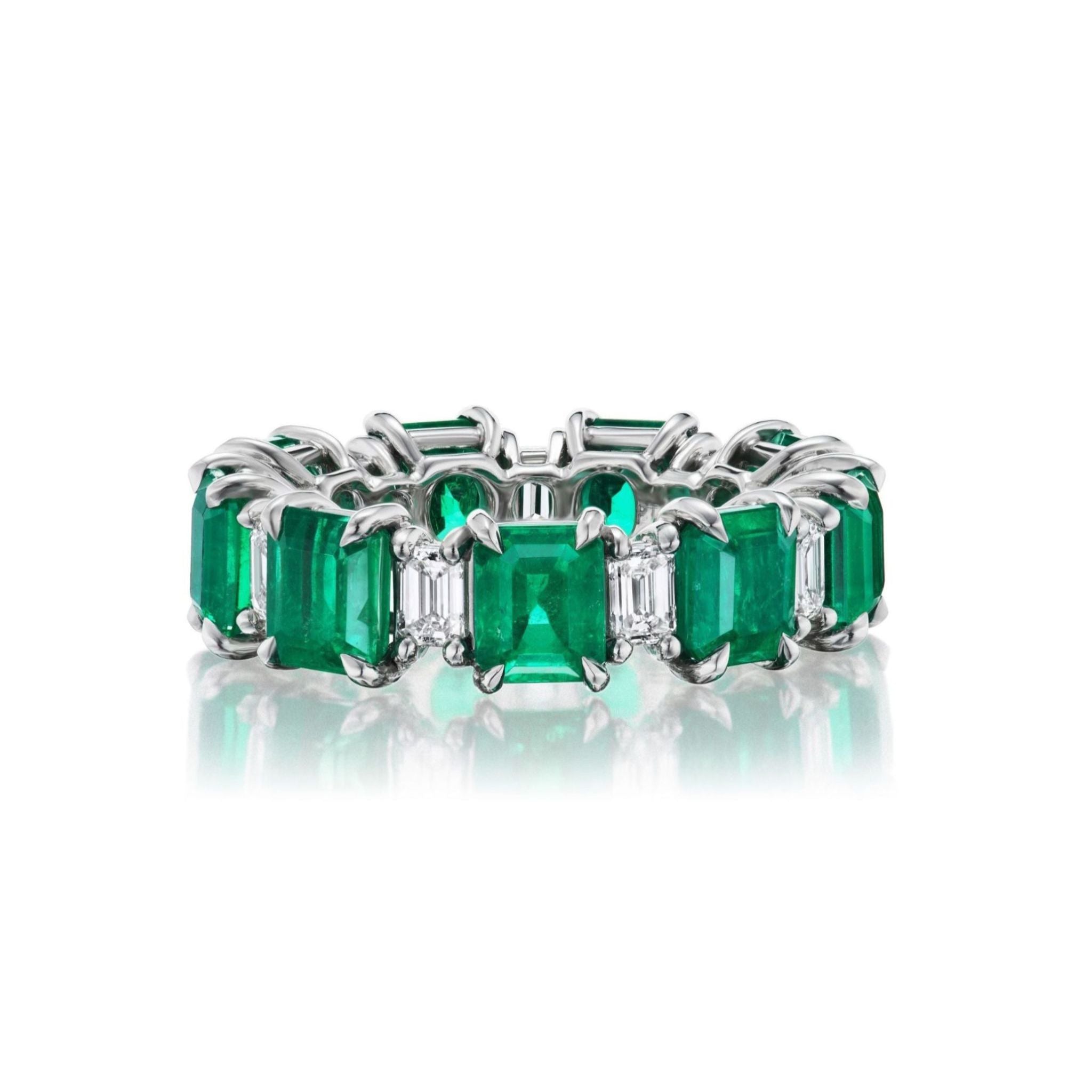 The Alternating Emerald Cut Diamond and Emerald Eternity Band showcases a seamless platinum design with alternating emerald-cut green emeralds and emerald-cut diamonds, elegantly displayed on a reflective white surface.