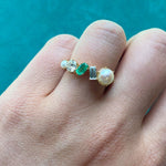 A hand wearing the Mixed Shape Pearl, Diamond and Emerald Half-Eternity Band, featuring a bespoke design with pearl, emerald, diamond, rectangular gem, and a small round stone accents, is displayed against a teal background.