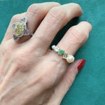 A hand with red nail polish wears two rings—one striking engagement ring with a large yellow stone and pink accents, and the Mixed Shape Pearl, Diamond and Emerald Half-Eternity Band, featuring pearls, a green stone, and diamonds—against a teal background.