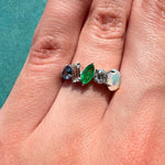 A hand adorned with the Mixed Shape Opal, Emerald, Diamond, and Sapphire Half-Eternity Band—crafted in 18k white gold and featuring five uniquely shaped gemstones in shades of blue sapphire, clear diamond, green emerald, white diamond, and a luminous oval opal—rests elegantly against a teal background.