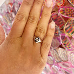 A hand with neatly manicured nails wears the Signet Ring with Burnish Set Hexagon Diamond, showcasing its hexagonal setting and sparkling diamond. Rows of colorful, patterned sunglasses form the backdrop.