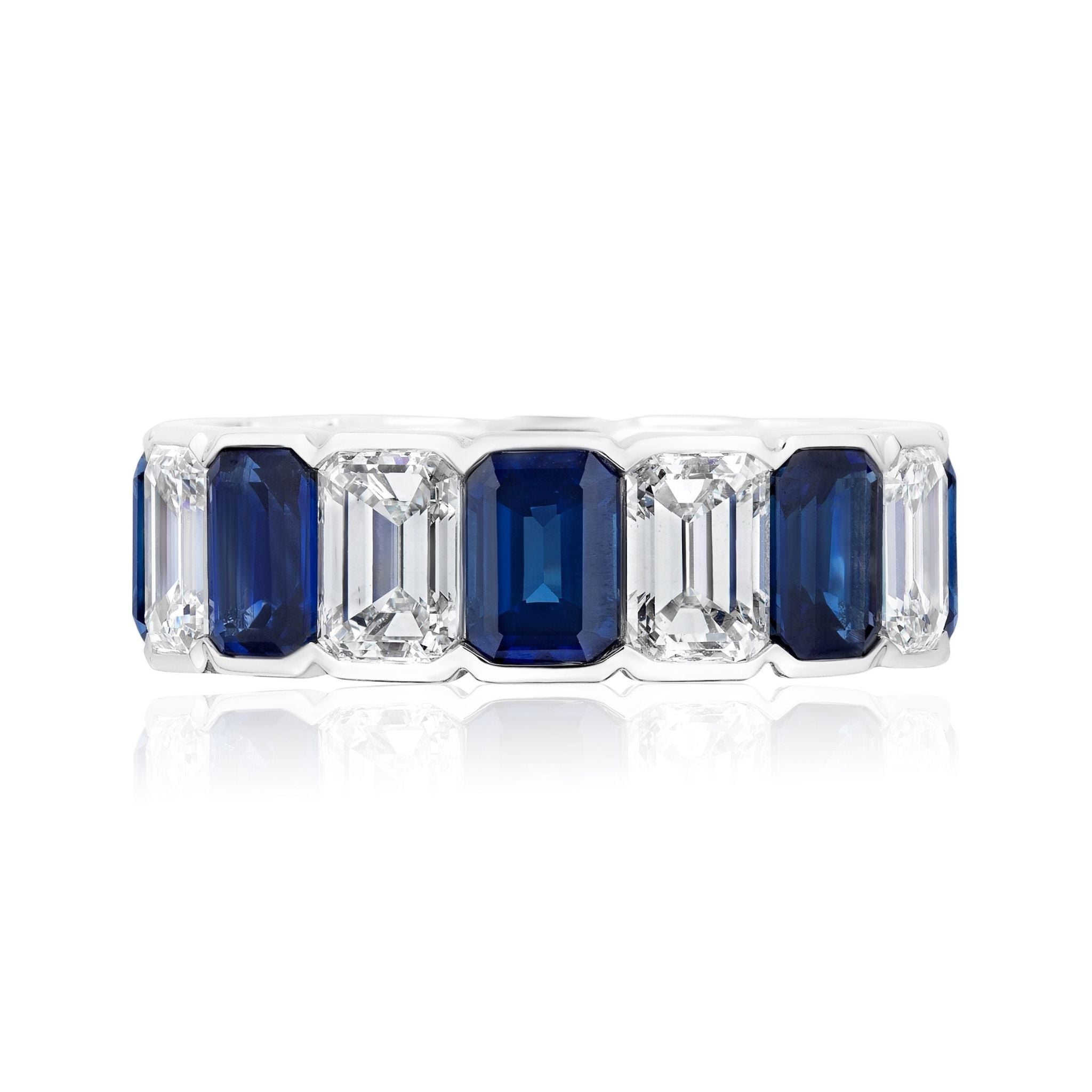 The Alternating Half-Bezel Emerald Cut Diamond and Sapphire Eternity Band is a white gold ring with alternating emerald-cut blue sapphires and white diamonds in a symmetrical design, displayed on a white background.