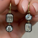 A hand holds Collet Set Emerald Cut and Round Diamond Drop Earrings in Yellow Gold and Black Rhodium, each earring featuring one emerald-cut and one round diamond. Fingernails are neatly manicured with light pink polish.
