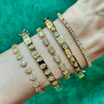 A wrist wears five Burnish Set Alternating Pear Shape Diamond Bangles in 18K yellow gold, each sparkling with clear SI+ diamonds, shown on a green textured background.