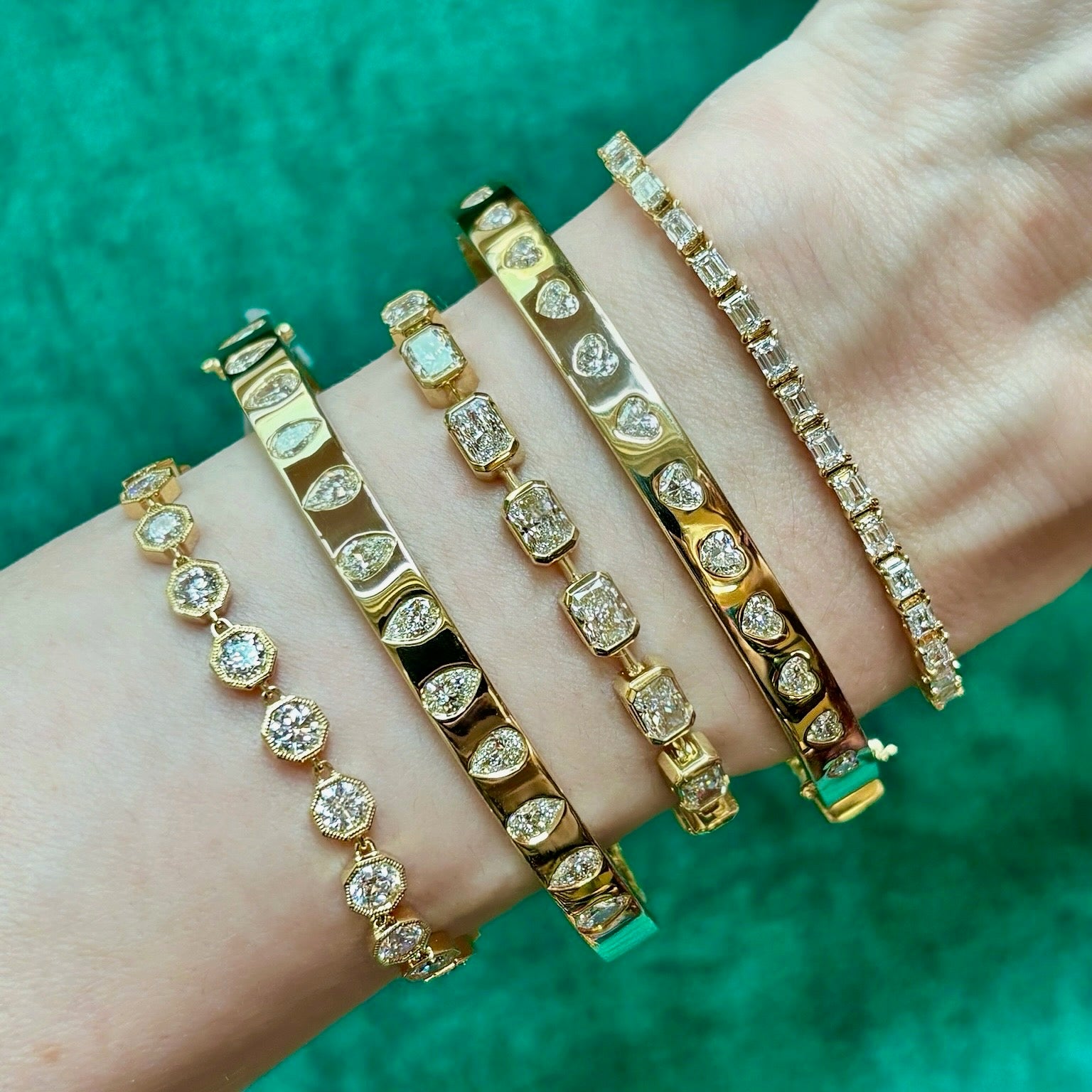A wrist wears five Burnish Set Alternating Pear Shape Diamond Bangles in 18K yellow gold, each sparkling with clear SI+ diamonds, shown on a green textured background.