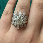 A hand wears a large, ornate diamond ring with a round center stone and starburst design of marquise, pear, and baguette diamonds atop the Confetti Diamond Ring Jacket in white and yellow gold, set against dark green fabric.