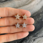 A hand holds two pairs of earrings; the top features gold star studs with sparkling stones, while the bottom pair is the Celestial Baby Star Studs in White Gold with Kite Diamonds, highlighting a celestial jewelry style.