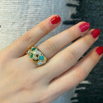 A hand with red nails wears the Bezel Set Cigar Band in Yellow Gold with Mixed Shape Diamonds and Emeralds, shown against a textured gray and white fabric background.