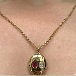 A person wears the Burnish Set Pear Shape Ruby and Emerald Cut Diamond Necklace, featuring an oval yellow gold pendant with a pear-shaped ruby and square emerald cut diamond.