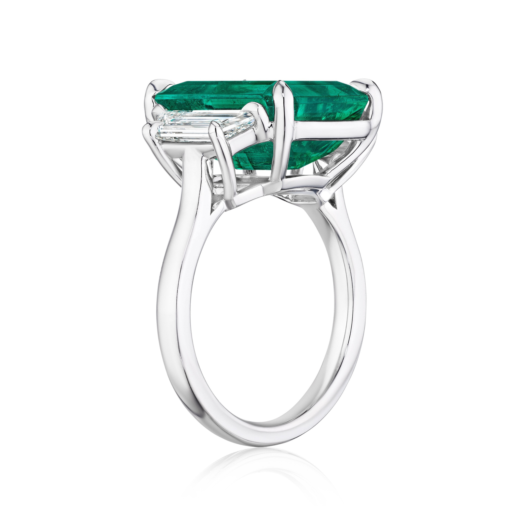 A Bespoke Emerald Cut Colombian Emerald Engagement Ring in White Gold features a prong-set rectangular green emerald with trapezoid baguette side stones, showcasing the gem’s depth and band’s intricate metalwork from the side view.