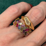A hand with two gold rings: one is the Geometric Burnish Set Ring in Yellow Gold with a shield cut diamond, while the other has zigzag rows of colorful rectangular gemstones. The background is dark green fabric.
