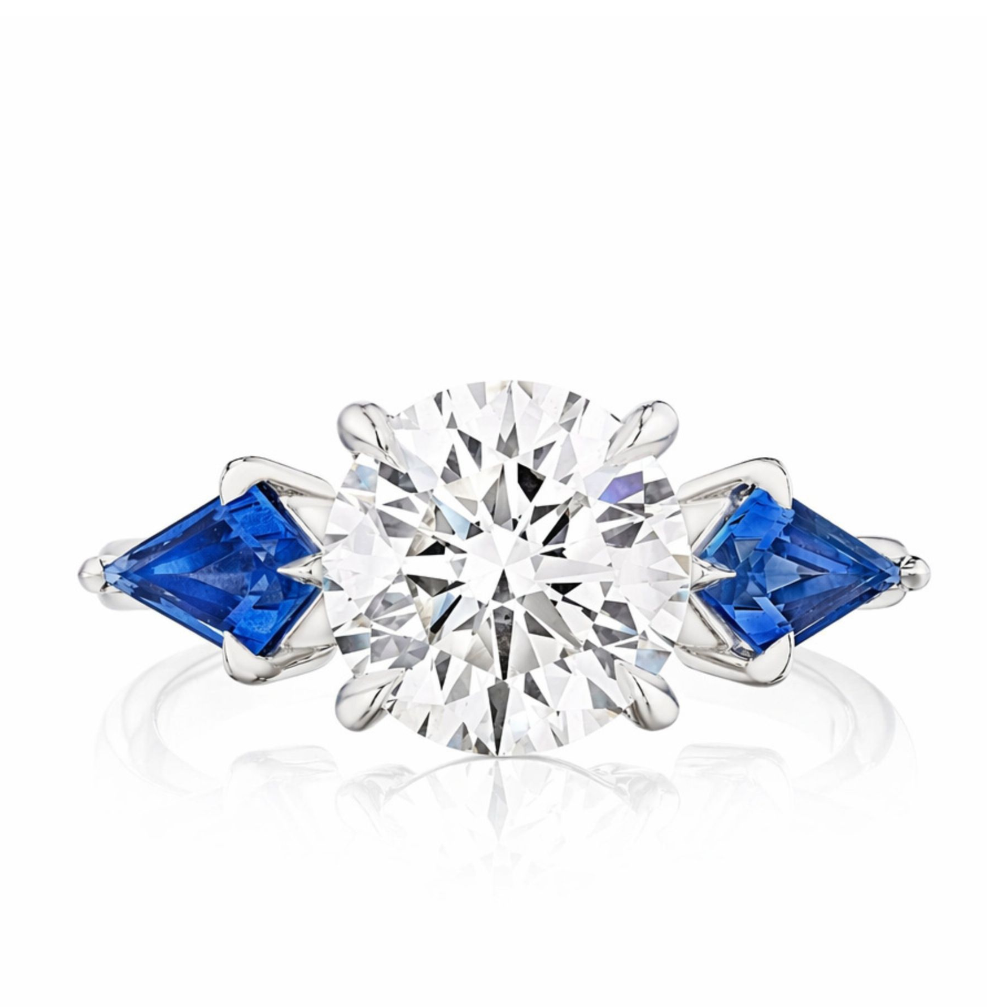The Bespoke Round Diamond Engagement Ring in White Gold features a round diamond centerpiece with kite-shaped blue sapphire side stones on each side.