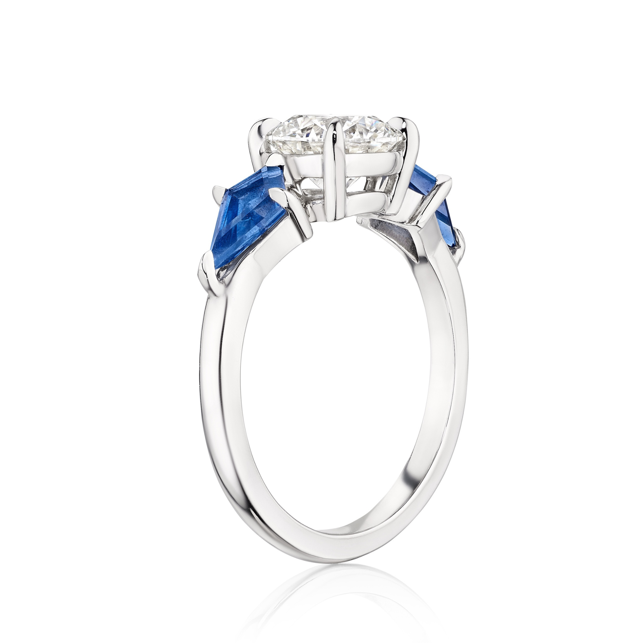 A close-up of the Bespoke Round Diamond Engagement Ring in white gold, showcasing a central round diamond with kite-shaped blue sapphire side stones in a prong setting, displayed against a white background.