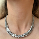 A close-up of a woman's neck showcases the Bezel Set Chevron Diamond Tennis Necklace in White Gold, featuring interlocking links in a geometric pattern. The necklace lies flat against her skin, with her blonde hair partially visible.