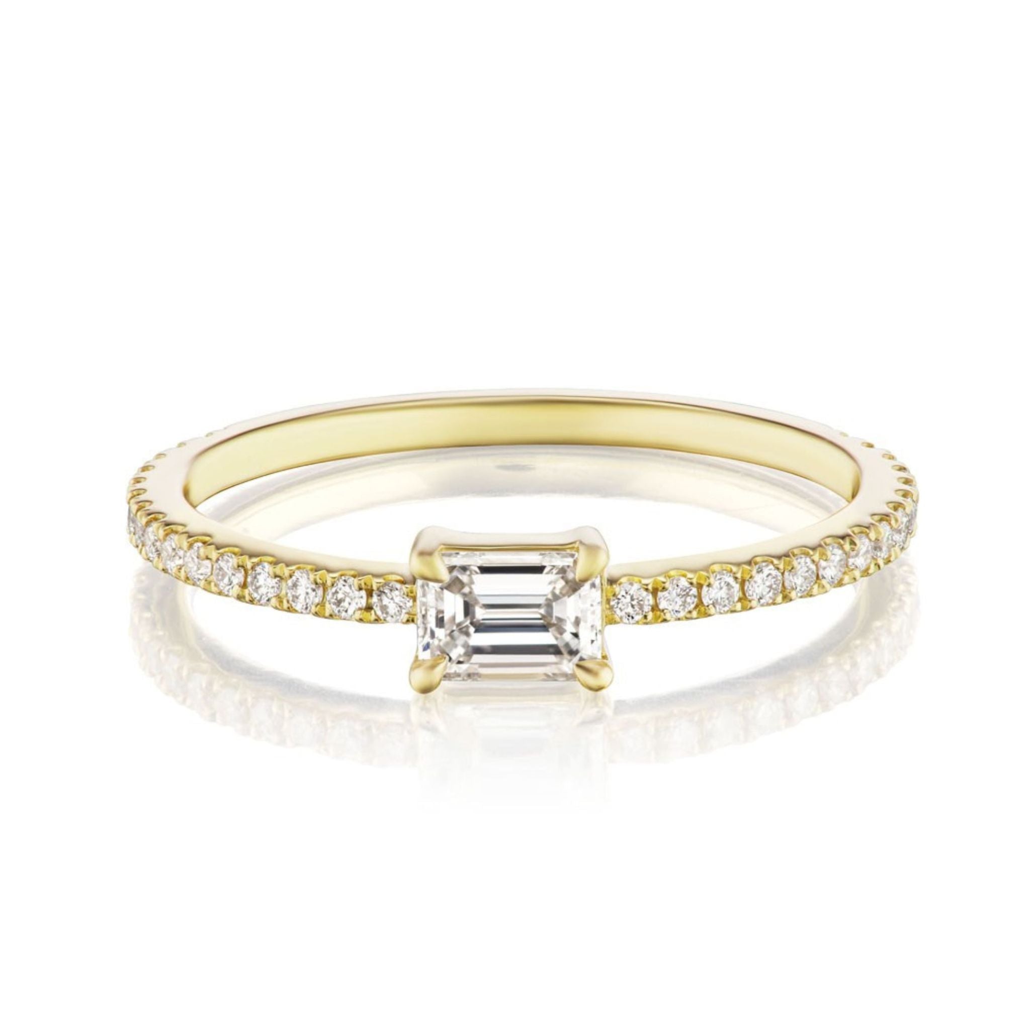 The East-West Set Emerald Cut Diamond Ring with Pavé Band in yellow gold features a striking emerald-cut diamond at the center, accented by pavé-set round diamonds on a slim band, displayed on a white background with subtle reflection.