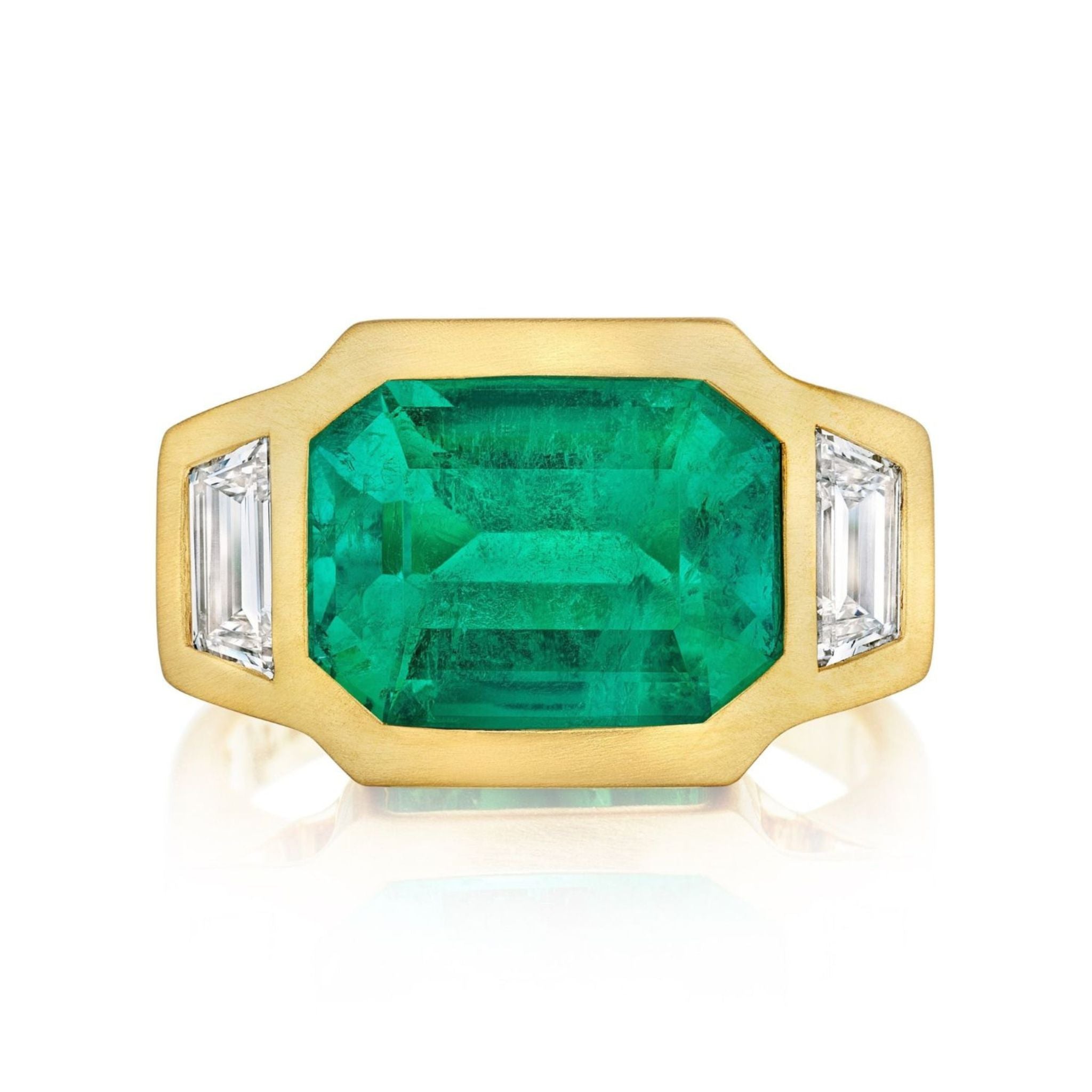The Bezel Set East-West Emerald Cut Emerald Three Stone Ring showcases a central emerald with trapezoid diamond side stones, all set in luxurious matte yellow gold.