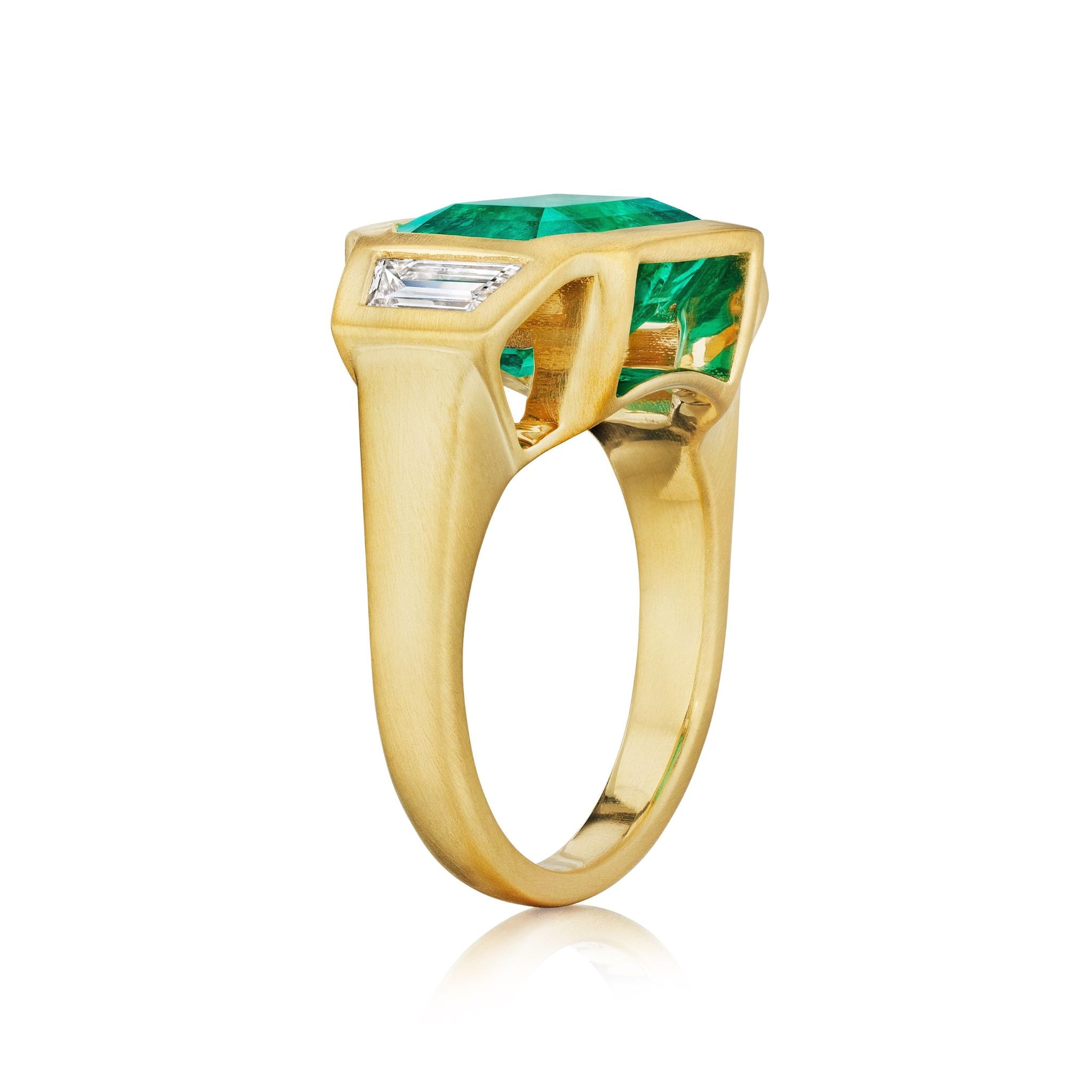 The Bezel Set East-West Emerald Cut Emerald Three Stone Ring features trapezoid diamond side stones set in matte yellow gold, showcased against a white background.