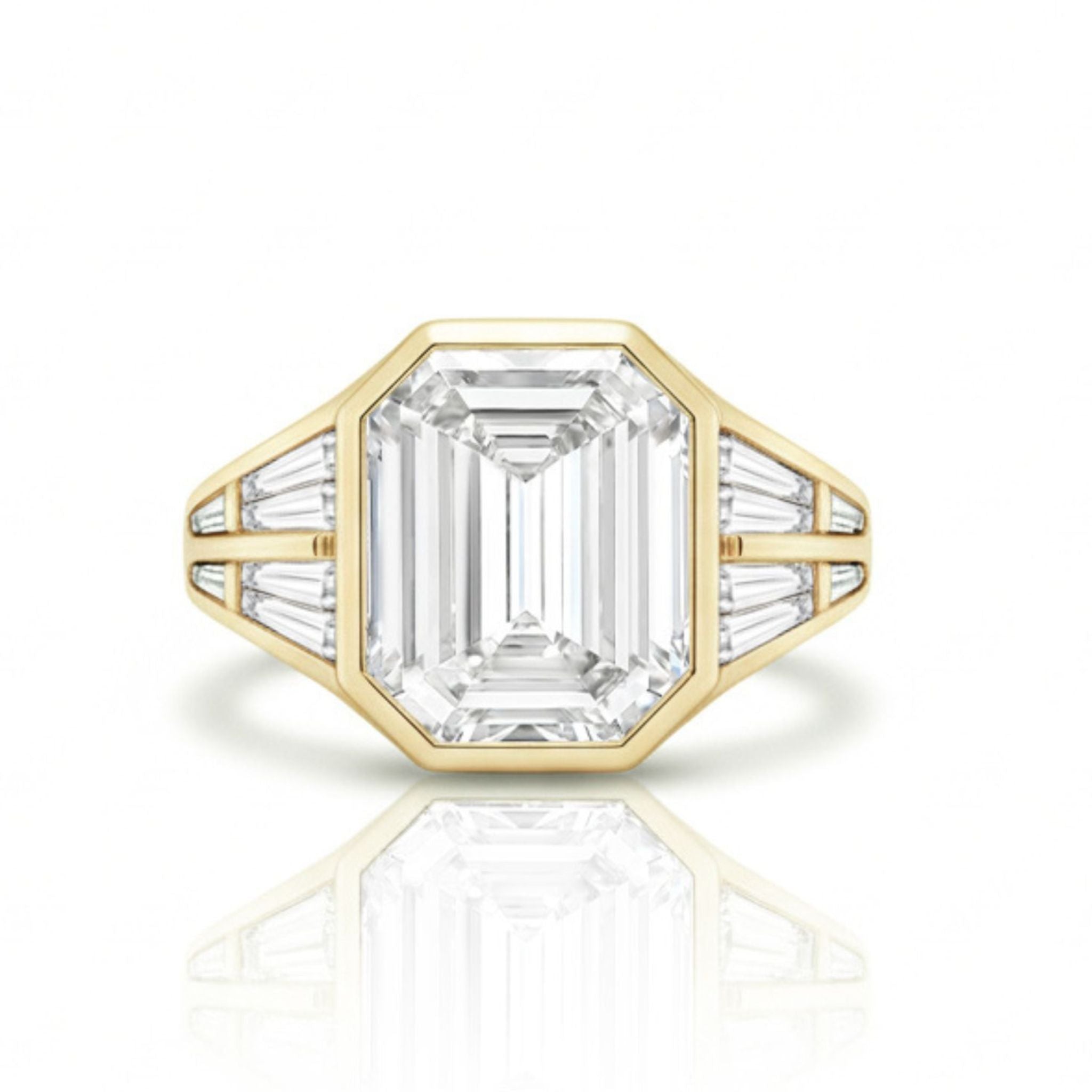 The Bezel Set Emerald Cut Diamond Engagement Ring with Tapered Baguette Side Stones in Yellow Gold is shown on a white background with a subtle reflection below.
