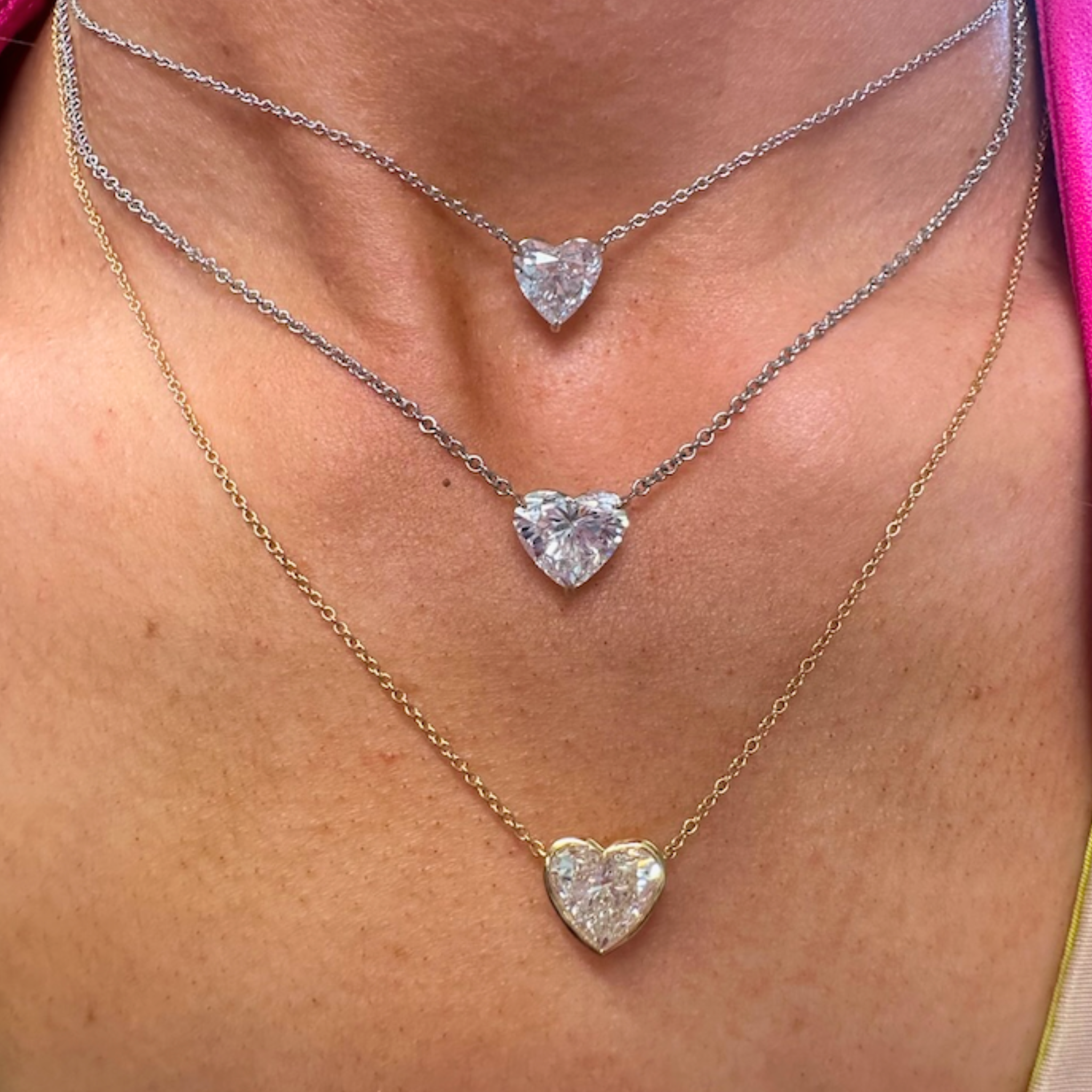 Close-up of a person wearing three Bezel Set Heart Shape Diamond Pendant Necklaces on delicate silver and 18K yellow gold chains, each sparkling elegantly against bare skin.