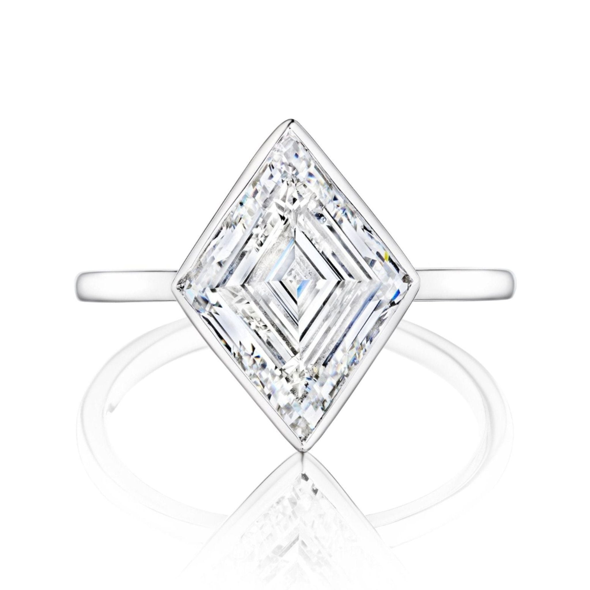 The Bezel Set Lozenge Diamond Engagement Ring, featuring a large diamond set at an angle on a thin 18K white gold band, is showcased against a white background with a soft reflection below.