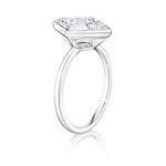The Bezel Set Lozenge Diamond Engagement Ring features a large emerald-cut diamond in a minimalist, thin 18K white gold band, showcased against a white background.
