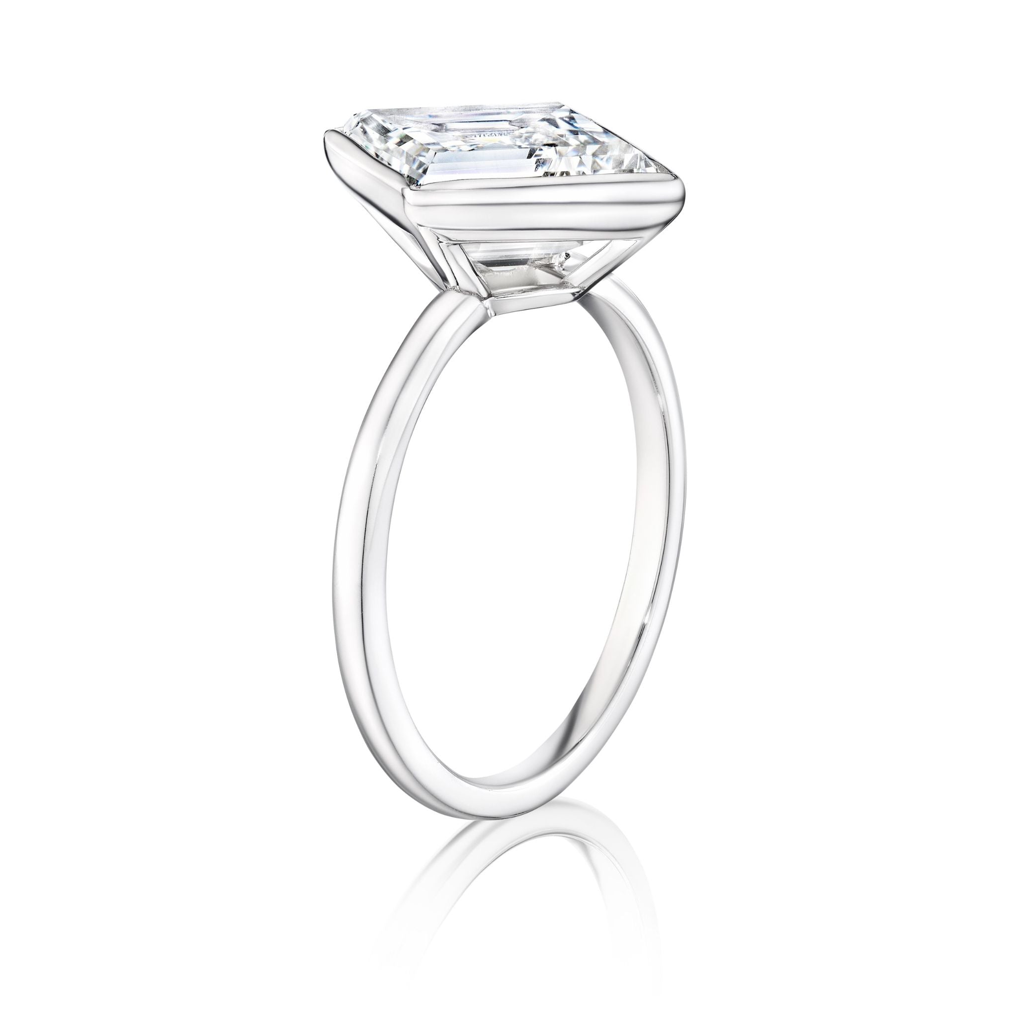 The Bezel Set Lozenge Diamond Engagement Ring features a large emerald-cut diamond in a minimalist, thin 18K white gold band, showcased against a white background.