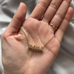 A hand with a gold ring holds the Bezel Set "Mama" Diamond Pendant Chain Necklace in Yellow Gold, featuring large decorative letters. The background is soft gray fabric.