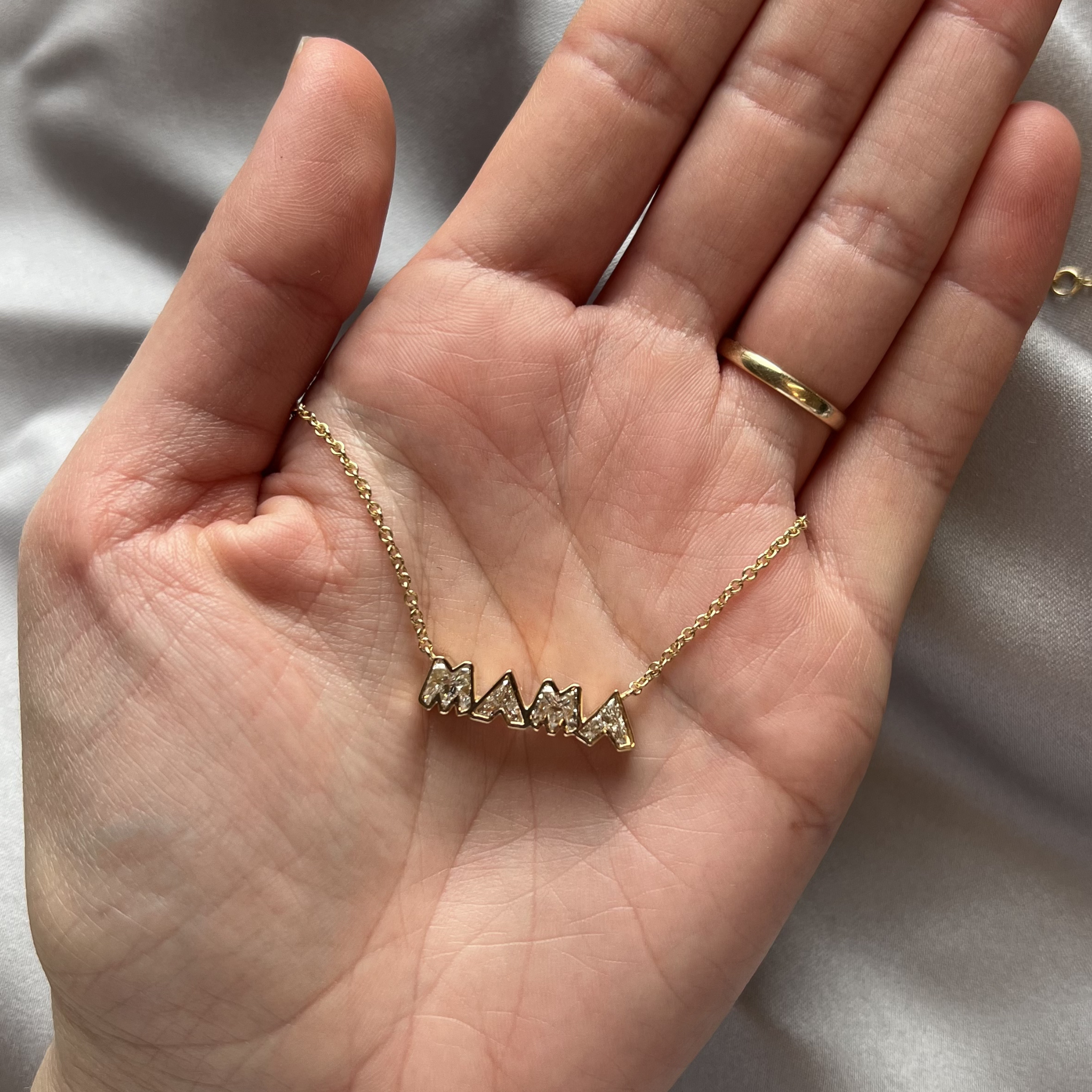 A hand with a gold ring holds the Bezel Set "Mama" Diamond Pendant Chain Necklace in Yellow Gold, featuring large decorative letters. The background is soft gray fabric.