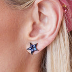 Close-up of an ear wearing Celestial Star Studs in Black Rhodium with kite sapphires; blonde hair and a pink hair accessory are partially visible, highlighting the celestial jewelry style.