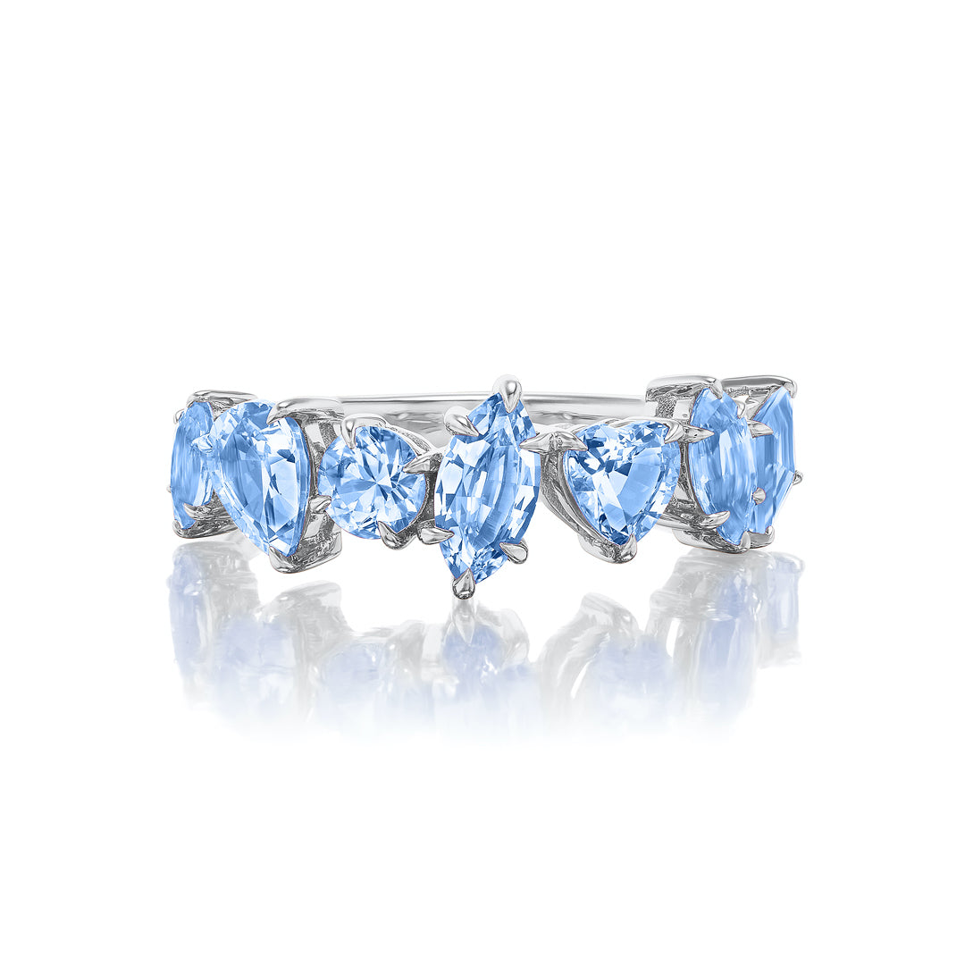 The Sapphire Mixed Shape Cluster Ring features five light blue sapphires—oval, round, marquise, pear, and heart—set in a silver half eternity band and displayed on a reflective white surface.
