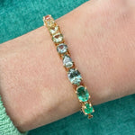 A Mixed Shape Diamond and Multicolor Sapphire Tennis Bracelet featuring vibrant green, blue, and yellow sapphires is worn on the wrist, with a green top visible in the background.