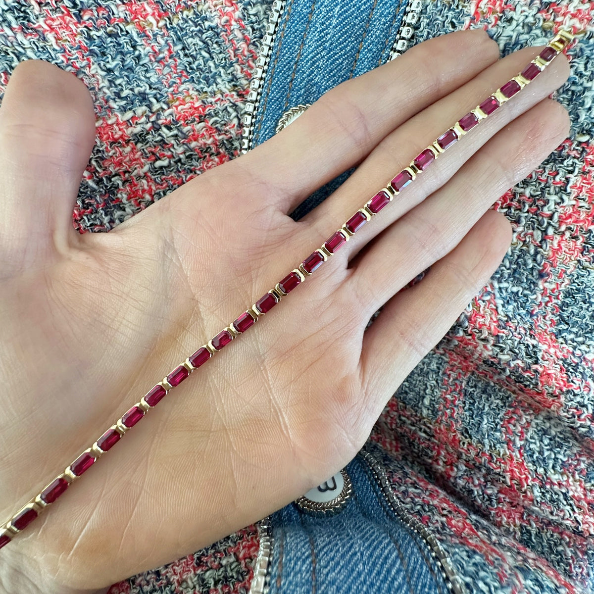 A hand holds the East-West Emerald Cut Ruby Tennis Bracelet in 18k yellow gold over textured fabric with pink, white, blue threads and soft blue denim.