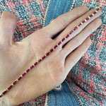 A hand holds the East-West Emerald Cut Ruby Tennis Bracelet in 18k yellow gold over textured fabric with pink, white, blue threads and soft blue denim.