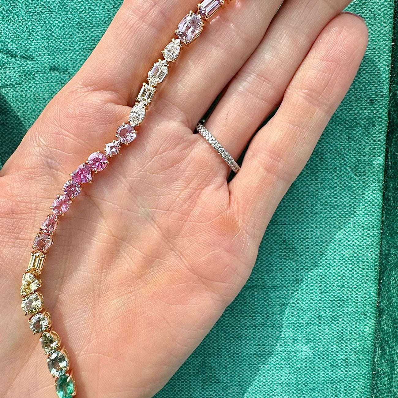 A hand with a silver diamond ring holds the Mixed Shape Diamond and Multicolor Sapphire Tennis Bracelet, adorned with pink, yellow, green, and clear diamonds and sapphires, set against a turquoise fabric background.